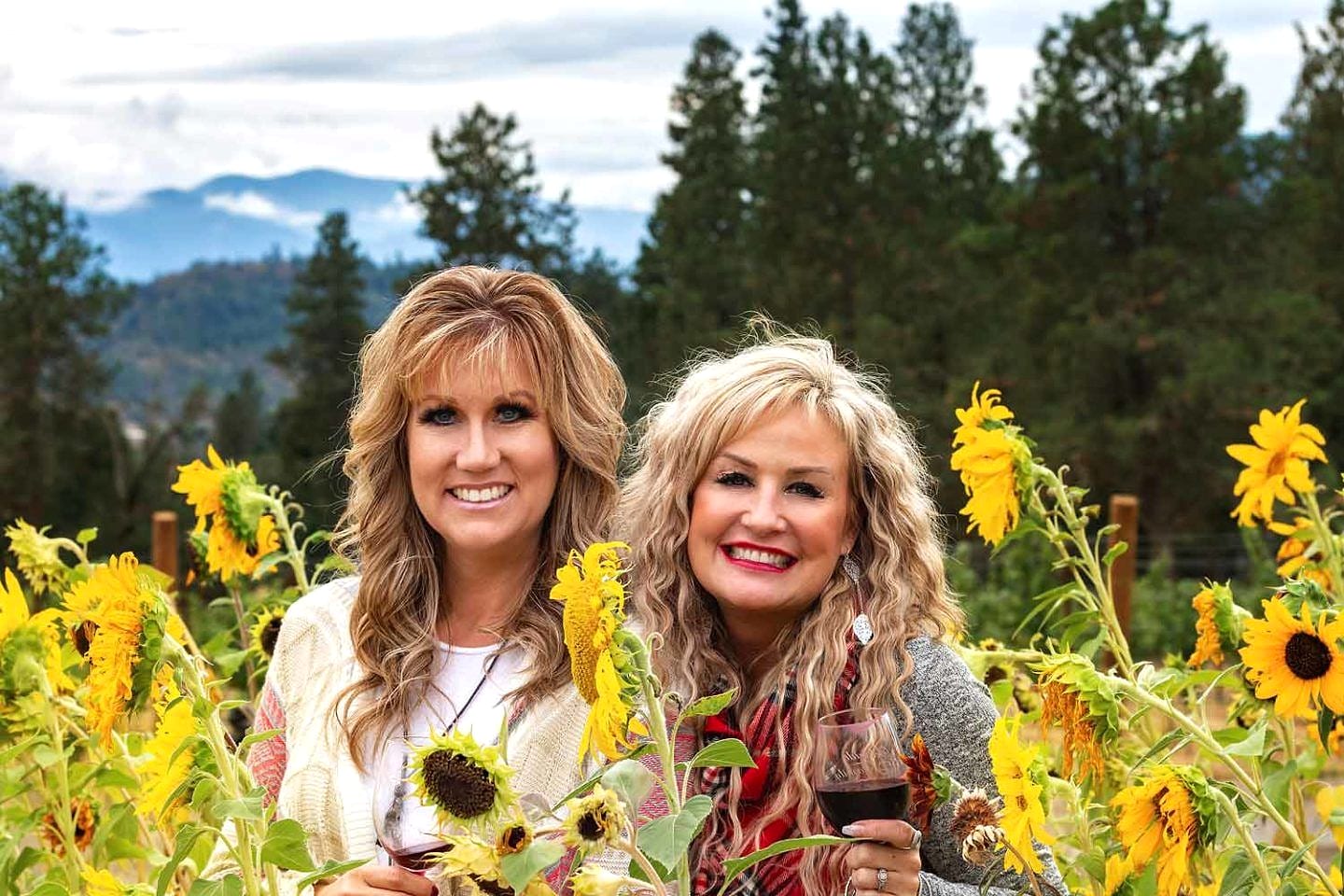 Two Salon Sisters holding wine glasses in a sunflower field with mountains behind